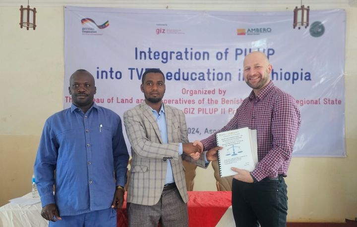 Three man standing in front of a banner that says "Integration of PILUP into TVET education in Ethipia", shaking hands as one man hands over the desing of the new training modules.