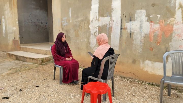 Two women sitting in front of each other on simple plastic chairs outdoors in front of a grey wall, one of them has a smartphone in her hands, used for the data collection of the project.