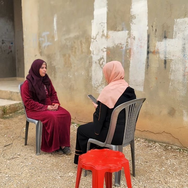 Two women sitting in front of each other on simple plastic chairs outdoors in front of a grey wall, one of them has a smartphone in her hands, used for the data collection of the project.