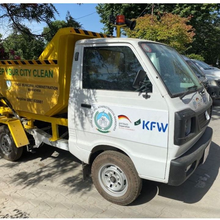 A public waste collection area with the project sign and a garbage truck with the project and donor's logo.